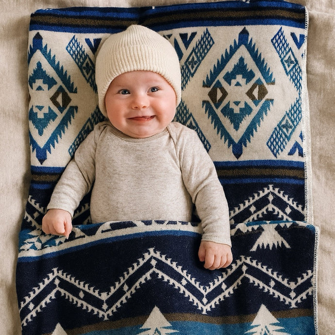 Smiling baby wrapped in a soft blue and cream Aztec-patterned Alpaca Threadz blanket, sitting in a cozy neutral nursery with natural light and warm textures.